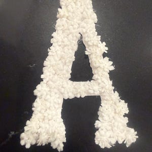 May include: A capital letter A shaped decoration covered in small, white, multi-petaled floral embellishments. The structure is entirely white, featuring a dense arrangement of individual flower shapes that create a textured, three-dimensional surface.