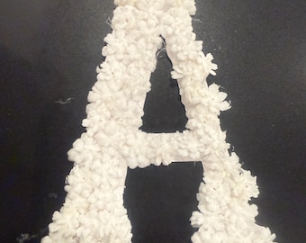 May include: A capital letter A shaped decoration covered in small, white, multi-petaled floral embellishments. The structure is entirely white, featuring a dense arrangement of individual flower shapes that create a textured, three-dimensional surface.