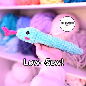 Può includere: Un serpente di peluche all'uncinetto color azzurro con lingua rosa, occhi neri e guance rosa. L'immagine include il testo PDF PATTERN ONLY e il testo LOW-SEW! in lettere bianche con contorno nero, indicando un modello per uncinetto.