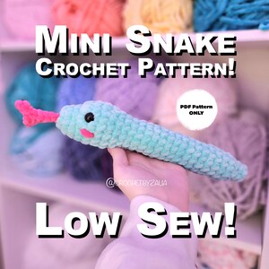 May include: A crocheted mini snake pattern in the image. The snake is light blue with a pink tongue and black eyes. The text "Mini Snake Crochet Pattern!" and "Low Sew!" are displayed. The background features colorful yarn.
