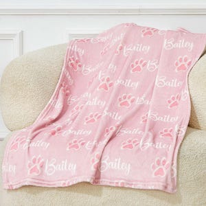May include: A pink blanket with a repeating pattern of paw prints and the name "Bailey" in white. The blanket is draped over a beige armchair, creating a cozy and inviting scene. The blanket is a soft, plush material.