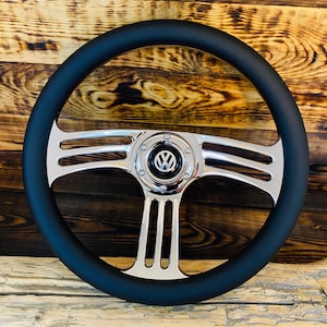 Könnte beinhalten: Ein schwarzes Lenkrad mit verchromter Mitte und Speichen. In der Mitte befindet sich ein Volkswagen-Logo. Das Lenkrad hat ein klassisches Design mit einem gepolsterten schwarzen Rand und einer polierten Chromoberfläche.