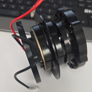 Könnte beinhalten: Ein schwarzer Lenkrad-Schnellverschluss-Adapter aus Metall mit kreisförmigem Design und einem inneren Ring in Goldoptik. Das Gerät verfügt über ein rotes und ein schwarzes Kabel, die seitlich herausragen, sowie ein mehrteiliges Befestigungsmuster.