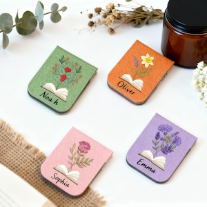 May include: Four felt bookmarks in green, orange, pink, and purple, each with a floral design and a name: Noah, Oliver, Sophia, and Emma. Each bookmark features an open book illustration. Ideal for book lovers.