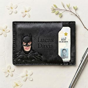 May include: A black leather card holder featuring an embossed Batman mask with a human face cutout, the name Lucas Davis engraved in the center, and a slot containing an identification card with a gold star and the text Class D Boat 05-20-2014.