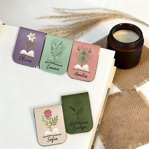 May include: Five floral magnetic bookmarks in shades of purple, green, pink, beige, and olive green. Each bookmark displays a unique floral design, an open book illustration, and a name. A brown candle is visible in the background.