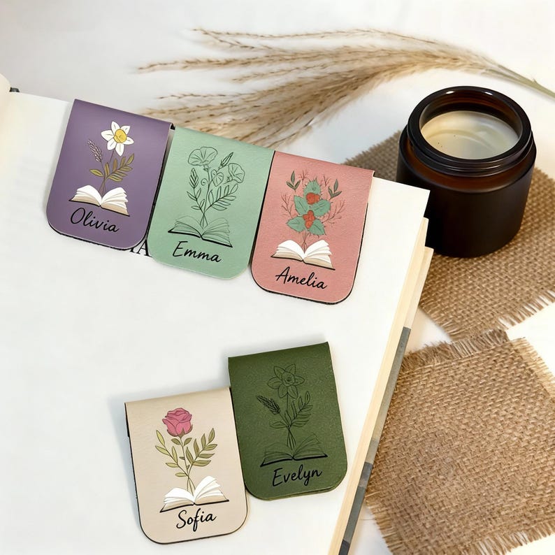 May include: Five floral magnetic bookmarks in shades of purple, green, pink, beige, and olive green. Each bookmark displays a unique floral design, an open book illustration, and a name. A brown candle is visible in the background.