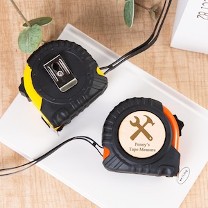 Könnte beinhalten: Zwei schwarz-gelbe Maßbänder mit Metallgürtelclips und schwarzen Zugbändern. Ein Maßband hat einen Holzring mit einem Hammer- und Schraubenschlüssel-Design und dem Text "Penny's Tape Measure". Die Maßbänder liegen auf einer weißen Oberfläche.