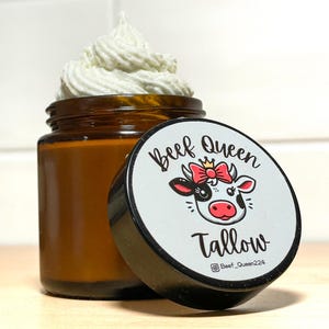 May include: A glass jar with a black lid containing a white whipped cream-like substance. The lid has a sticker with a cartoon cow wearing a pink bow and a crown. The sticker says "Beef Queen Tallow" and includes the Instagram handle "@Beef_Queen224".