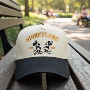 Może przedstawiać: Kremowa czapka bejsbolówka z czarnym daszkiem z napisem "DISNEYLAND" wyszytym złotą nicią. Poniżej znajdują się haftowane wizerunki Myszki Miki i Myszki Minnie, z napisem "EST. 1955". Czapka leży na drewnianej ławce.