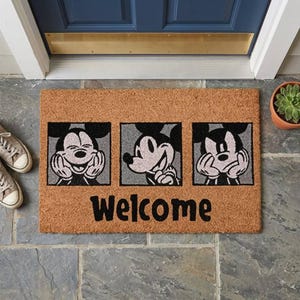 Peut inclure: Un paillasson rectangulaire en fibre de coco marron avec trois panneaux carrés, chacun présentant une illustration en noir et blanc de Mickey Mouse avec des expressions différentes. Le mot Welcome est écrit en grandes lettres noires.