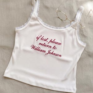 May include: White ribbed vest top with lace trim and the embroidered text "if lost, please return to William Johnson". Gold-rimmed glasses rest on the top. The vest top has thin straps.