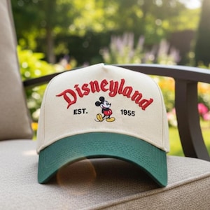 Może przedstawiać: Beżowa i zielona czapka bejsbolowa z napisem "Disneyland" w czerwonym skrypcie, wizerunkiem Myszki Miki i tekstem "EST. 1955". Czapka leży na krześle.