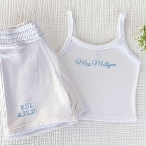 Puede incluir: Conjunto de pijama blanco a juego con camiseta sin mangas y pantalones cortos. La camiseta sin mangas tiene tirantes finos y el texto bordado "Mrs. Matiger" en azul claro. Los pantalones cortos tienen el texto bordado "EST. 8.22.25" en azul claro.