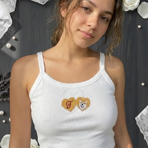 Puede incluir: Camiseta sin mangas blanca acanalada con bordado dorado en forma de corazón con la letra "T" y un perro Shiba Inu. La camiseta tiene tirantes finos y cuello redondo.