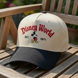May include: A beige and navy baseball cap with the words "Disney World" in red script, an image of Mickey Mouse, and the year "1971". The cap has a curved peak and a button on top. The hat is resting on a wooden chair.