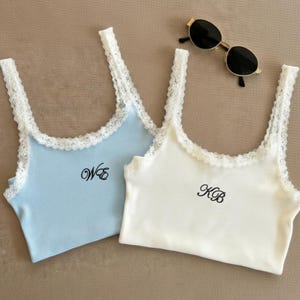 May include: Two ribbed tank tops, one light blue and one white, with white lace trim. Each top has black embroidered initials. A pair of black sunglasses with gold frames rests nearby.