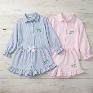 Op de afbeelding: Twee sets gestreepte pyjama's. De ene set is blauw en wit, de andere roze en wit. Elke set bevat een overhemd met knopen en een zak en een short met een ruche zoom. De initialen "XYZ" zijn op elk stuk geborduurd.