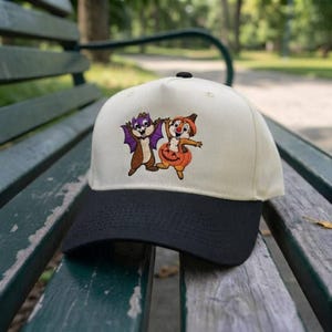 Peut inclure: Une casquette de baseball couleur crème avec une visière noire, ornée d'une broderie représentant deux tamias en costumes d'Halloween. L'un porte des ailes de chauve-souris violettes, l'autre est dans une citrouille orange. Calotte structurée.