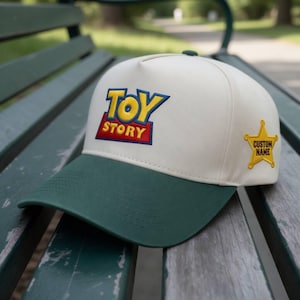 Può includere: Un cappellino da baseball con corona bianca e visiera verde. Il cappellino presenta il logo "Toy Story" in ricamo blu, giallo e rosso. Una toppa a forma di stella gialla con la scritta "CUSTOM NAME" è sul lato.