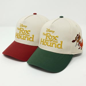 May include: Two baseball caps with the text "Disney The Fox and the Hound" embroidered in gold. One cap has a red bill and the other has a green bill. The caps have a cream-colored crown and a small button on top.