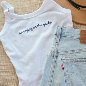 Puede incluir: Camiseta sin mangas blanca con la frase bordada "no crying on the yacht" en azul marino, combinada con shorts vaqueros de lavado claro. Un sombrero color canela es visible en el fondo.