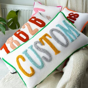 May include: White rectangular decorative pillows with colorful embroidered text. One pillow says "CUSTOM" in yellow, gray, and blue. Other pillows have orange and pink text. Green trim outlines the pillows.