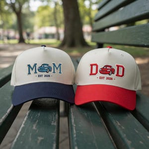 May include: Two baseball caps with embroidered designs. One cap has a cream-colored crown and navy blue brim, with the word "MOM" and a car graphic. The other cap has a cream-colored crown and red brim, with the word "DAD" and a car graphic. Both caps have "EST 2026" text.