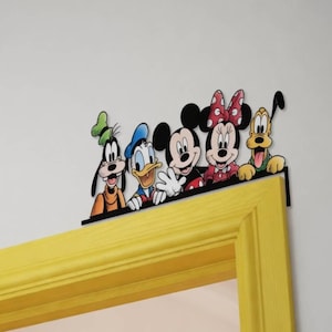 May include: A black metal door topper featuring a colorful illustration of Goofy, Donald Duck, Mickey Mouse, Minnie Mouse, and Pluto. The characters are depicted from the waist up, arranged in a row, and mounted on a flat black metal base.