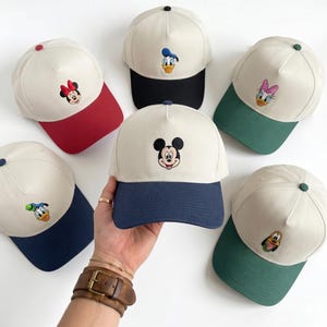 Op de afbeelding: Zes baseballpetten met gebroken witte kronen en gekleurde kleppen. Elke pet heeft een geborduurd karaktergezicht op het voorpaneel. De kleppen zijn rood, zwart, donkerblauw en groen. De karakters zijn onder andere Mickey Mouse, Minnie Mouse, Donald Duck, Daisy Duck en Pluto.