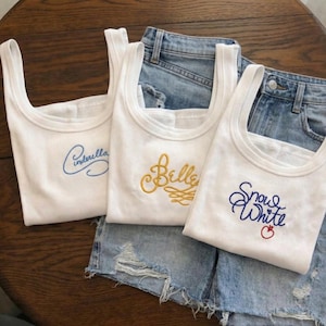 Peut inclure: Trois débardeurs blancs avec des noms brodés : "Cinderella" en bleu, "Belle" en or et "Blanche-Neige" en bleu avec une pomme rouge. Les hauts sont présentés sur un short en jean.