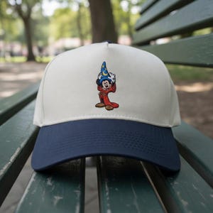 Op de afbeelding: Een baseballpet met een crèmekleurige kroon en een marineblauwe rand. De pet heeft een geborduurd beeld van Mickey Mouse in een tovenaarskostuum. De afbeelding toont Mickey Mouse die zijn tovenaars hoed vasthoudt. De hoed is blauw met een maan en sterren.