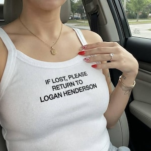 Könnte beinhalten: Ein weißes geripptes Tanktop mit schwarzem Text, der IF LOST, PLEASE RETURN TO LOGAN HENDERSON lautet. Das Kleidungsstück wird mit einer goldenen Halskette mit sechseckigem Anhänger, einem silbernen Ring mit quadratischem Stein und einem goldenen Armband getragen.