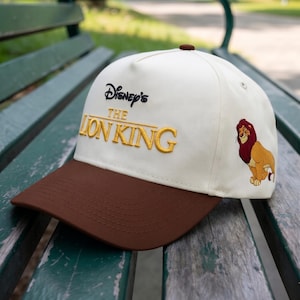May include: A baseball cap featuring the text "Disney's The Lion King" embroidered in gold thread. The cap has a brown bill and a cream-colored crown. A cartoon image of Simba is on the side.