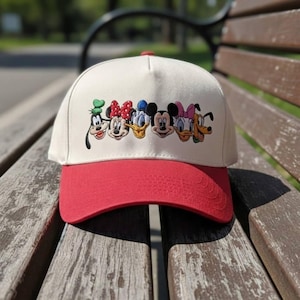 Może przedstawiać: Czapka bejsbolowa z kremową koroną i czerwonym daszkiem. Czapka posiada haftowany wzór postaci z kreskówek Disneya, w tym Myszki Miki i Kaczora Donalda.