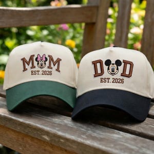 Może przedstawiać: Dwie czapki bejsbolowe z napisami "MOM" i "DAD" w brązowych literach blokowych, z motywami Myszki Miki i Minnie. Czapka "MOM" ma zielony daszek, a czapka "DAD" ma granatowy daszek. Obie czapki mają wyszyte "EST. 2026" poniżej.