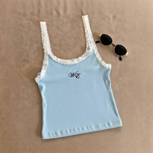 May include: A light blue tank top with white lace trim and straps. The top has the embroidered letters "WE" in black on the front. A pair of black sunglasses with gold frames rests on the side.