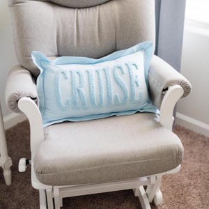 Op de afbeelding: Een lichtblauw sierkussen met het woord "CRUISE" geborduurd in een lichtere blauwe kleur, geplaatst op een grijs gestoffeerde schommelstoel. De stoel heeft witte houten armleuningen en poten, tegen een neutrale achtergrond.