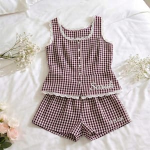 May include: A two-piece pajama set in a burgundy and white gingham pattern. The top is a sleeveless tank with a scalloped lace trim and button-up front. The shorts also have a scalloped lace trim. The name "Sarah" is embroidered on the top, and "9/8/26" is on the shorts.