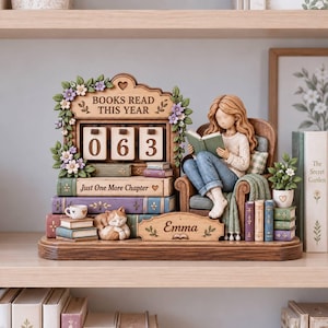 May include: A decorative wooden display featuring a counter with the text BOOKS READ THIS YEAR and the number 063. It includes a figurine of a person reading in a chair, a sleeping cat, stacks of books labeled Just One More Chapter, and a nameplate reading Emma, all adorned with floral accents.