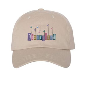 Pu&ograve; includere: Un cappello da baseball beige con un logo Disneyland colorato con bandiere e il testo "Disneyland".