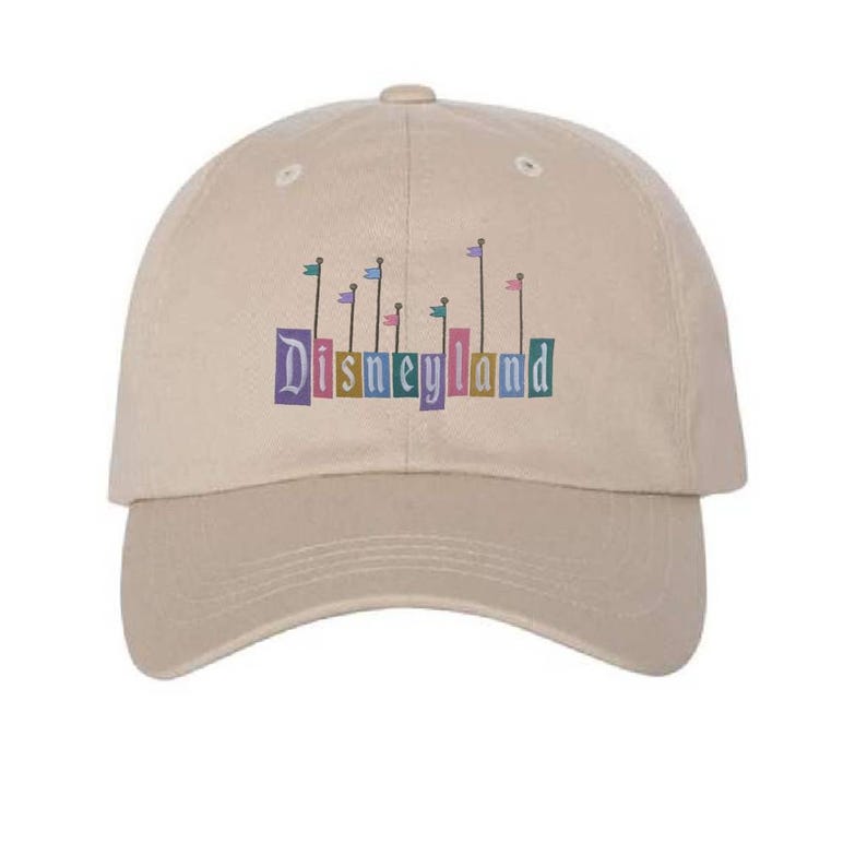 Pu&ograve; includere: Un cappello da baseball beige con un logo Disneyland colorato con bandiere e il testo "Disneyland".