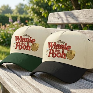 Puede incluir: Dos gorras de béisbol con el texto "Disney Winnie the Pooh" en rojo y amarillo. Una gorra tiene la visera verde, la otra negra. Ambas gorras tienen la copa beige y están sobre un banco de madera.