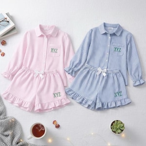 Op de afbeelding: Twee sets gestreepte pyjama's, één roze en één blauw, worden getoond. Elke set bevat een shirt met lange mouwen en knopen en een short met ruches. De shirts en shorts hebben de geborduurde letters "XYZ". Een witte strik siert elke short.