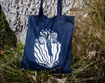 May include: A dark blue tote bag with white print. The print features a stylized tree with a person inside. The text "The Wild Swimmer" is printed below the tree.