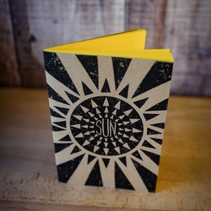 Puede incluir: Un pequeño cuaderno cerrado de color marrón con un diseño de sol en blanco y negro y la palabra "SUN" impresa en el centro. El cuaderno tiene una encuadernación amarilla.