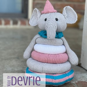 May include: A grey knitted elephant toy with a pink hat and a turquoise scarf. The elephant is a stacking toy with five rings, two pink, two white, and one grey. The toy is on a brown brick surface. Made by Devrie. Digital Download.