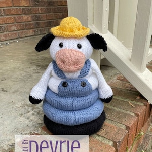 May include: Knitted toy cow wearing a yellow hat and blue overalls. The cow is a stacking toy with three rings. The toy is sitting on a brick surface. The text "MADE BY DEVRIE Digital Download" is on the image.
