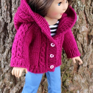 Puede incluir: Una chaqueta con capucha de punto magenta con botones blancos, usada por una mu&ntilde;eca. La chaqueta presenta un dise&ntilde;o de punto trenzado en las mangas y la parte delantera. La mu&ntilde;eca tambi&eacute;n lleva vaqueros azules y zapatos negros.