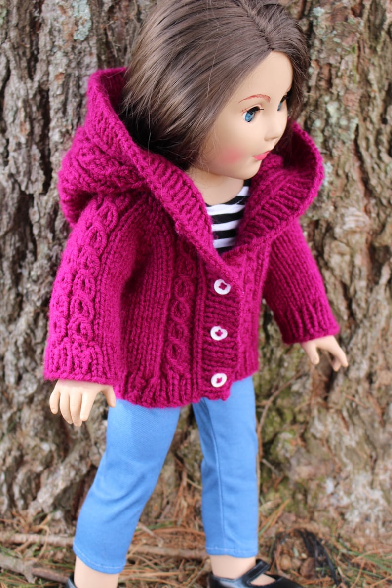 Puede incluir: Una chaqueta con capucha de punto magenta con botones blancos, usada por una mu&ntilde;eca. La chaqueta presenta un dise&ntilde;o de punto trenzado en las mangas y la parte delantera. La mu&ntilde;eca tambi&eacute;n lleva vaqueros azules y zapatos negros.